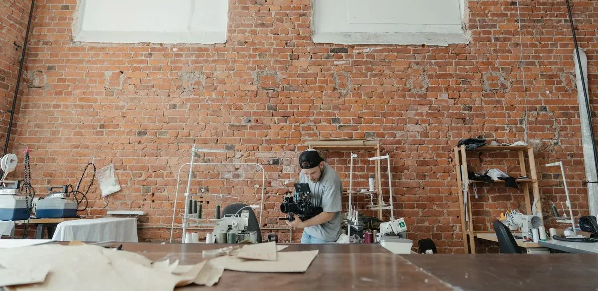 Videomaker filmando dentro de uma oficina de costura, entre máquinas e bancadas de trabalho