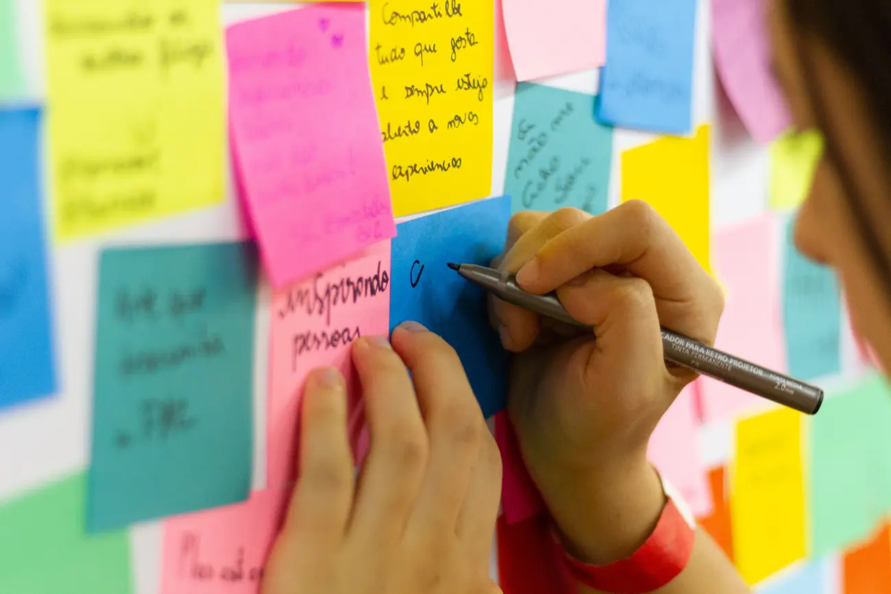 Mãos escrevendo em post-its coloridos colados em um mural durante o planejamento de conteúdo
