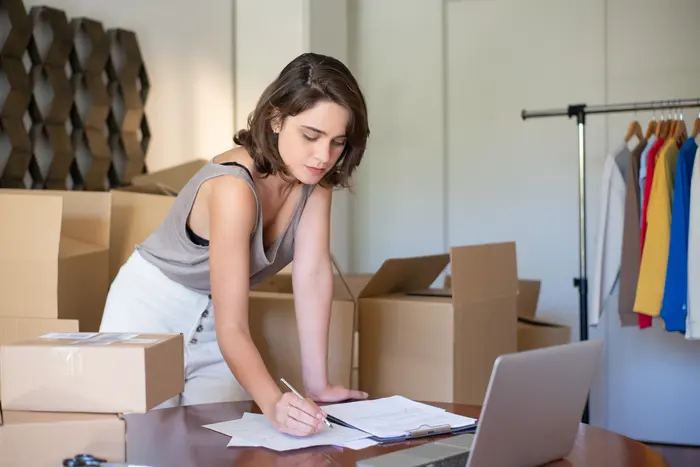 Dona de loja virtual conferindo pedidos entre caixas de papelão e o notebook