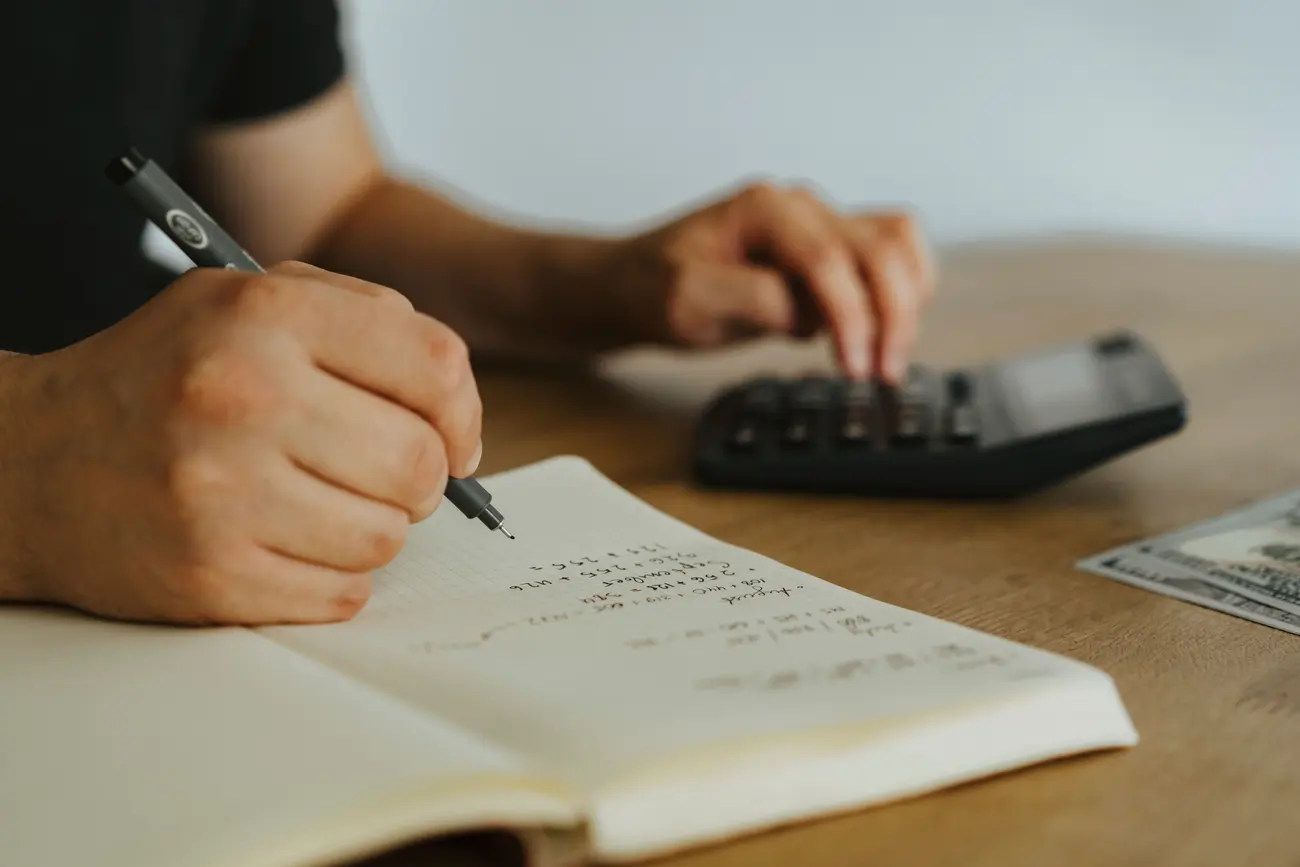 Pessoa escrevendo cálculos em um caderno ao lado de uma calculadora