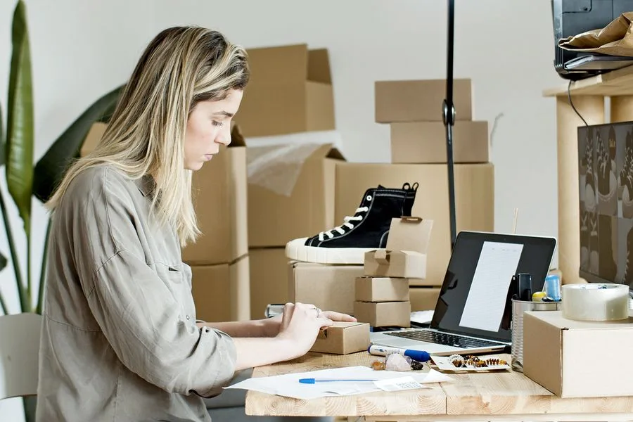 Empreendedora embalando pedidos da loja virtual em casa, com caixas e notebook na mesa