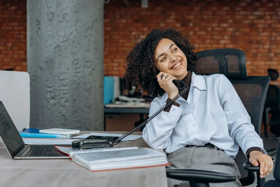 Atendente sorrindo ao telefone na mesa de trabalho, com notebook e agenda abertos
