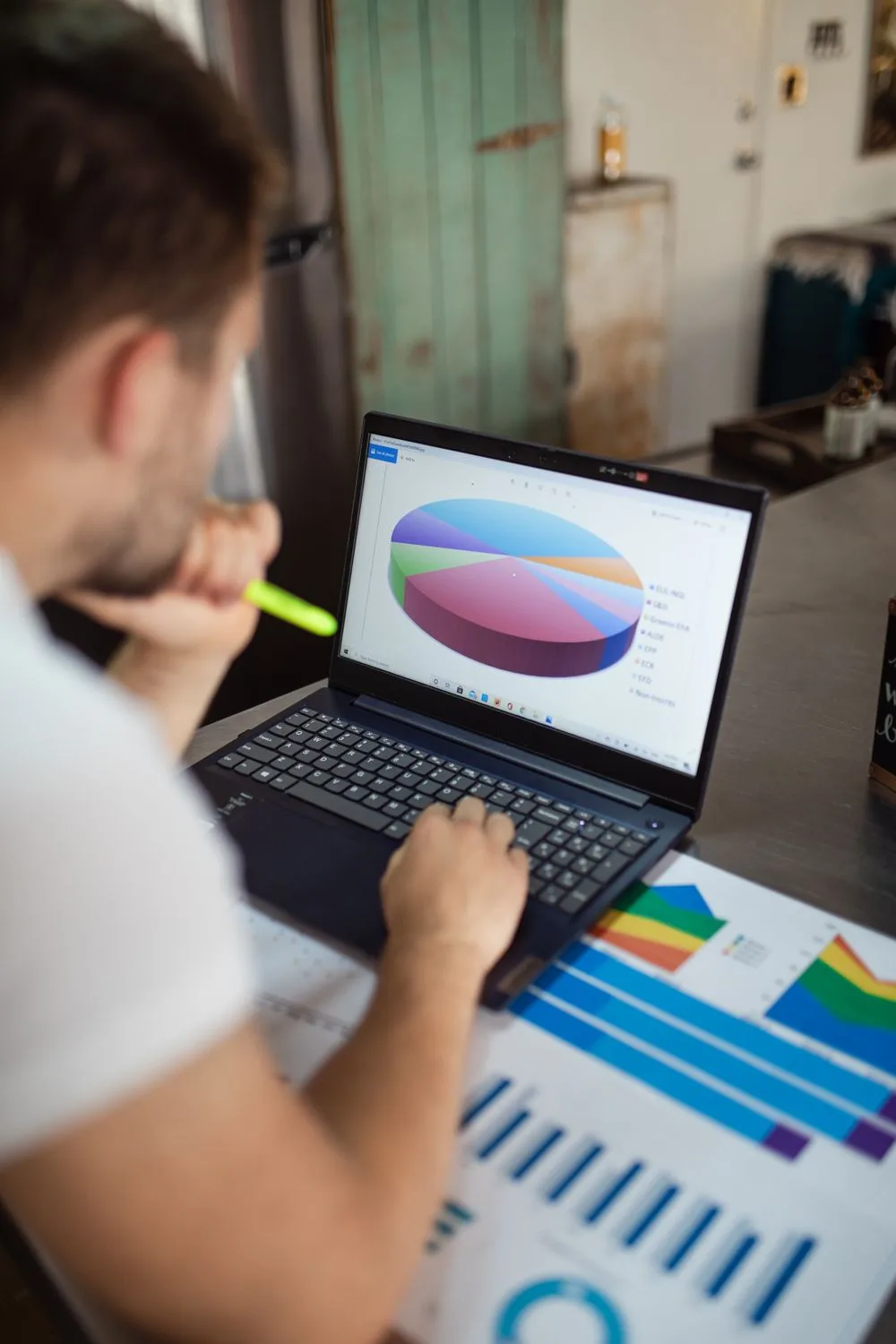Homem analisando um gráfico de desempenho na tela do notebook, com relatórios impressos sobre a mesa