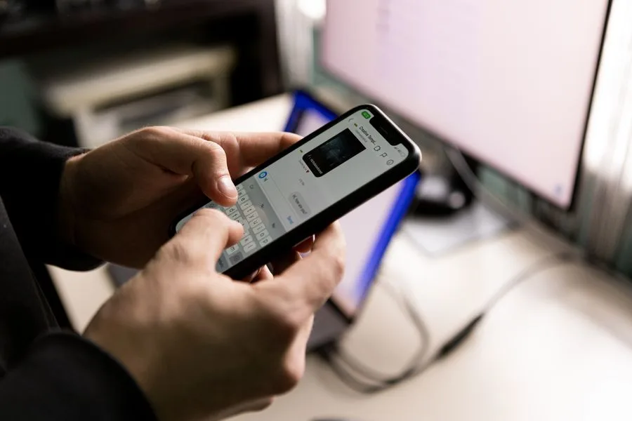 Pessoa digitando uma mensagem no celular em frente ao computador
