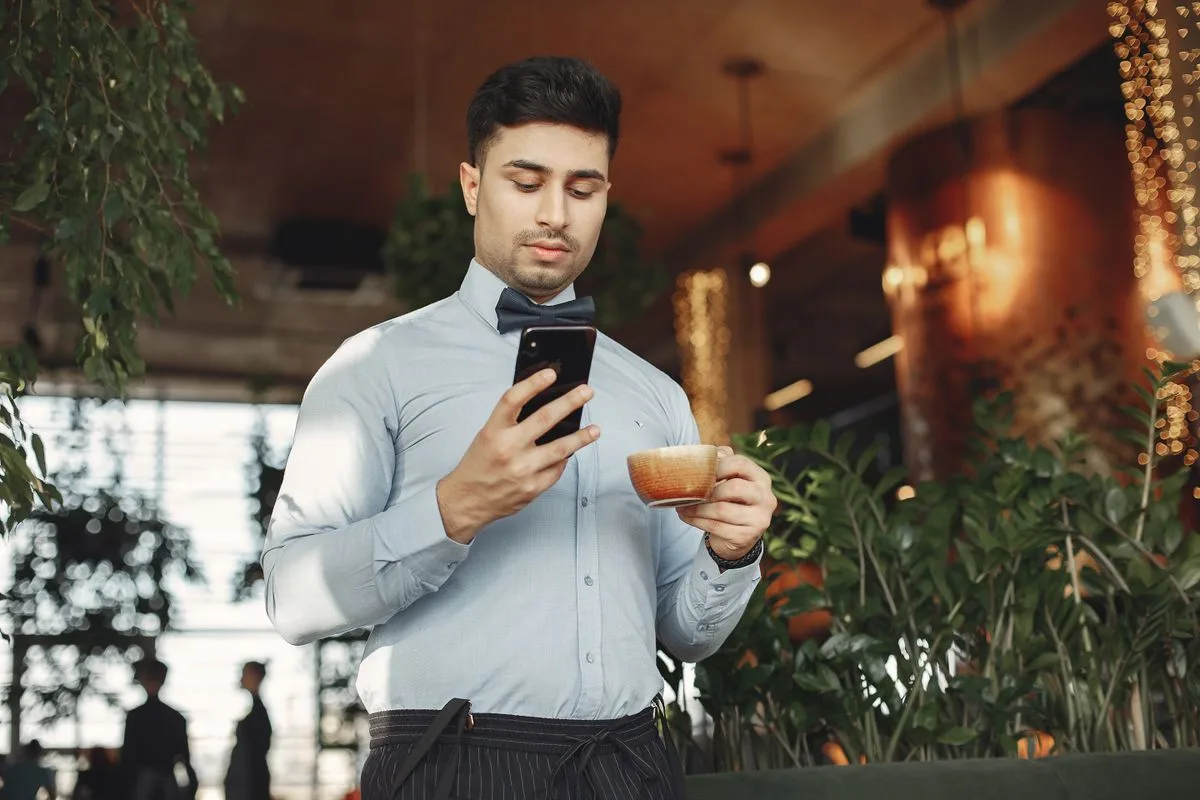 Homem em um café segurando uma xícara e olhando o celular