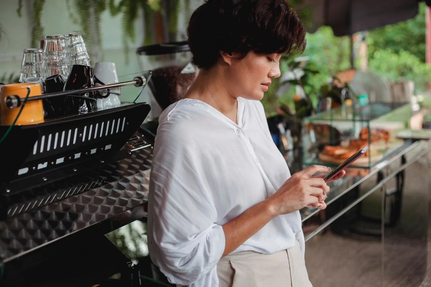 Dona de cafeteria encostada na máquina de café conferindo o celular